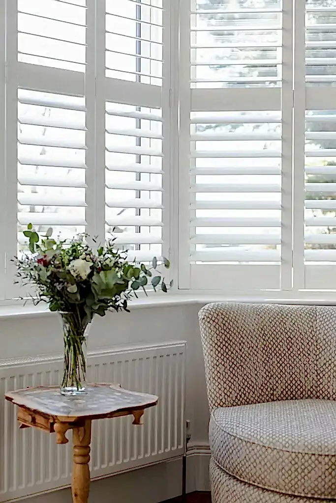 Plantation shutters in front of table with flowers and chair Plantation Shutters Project - Your #1 Choice for Shutters NI